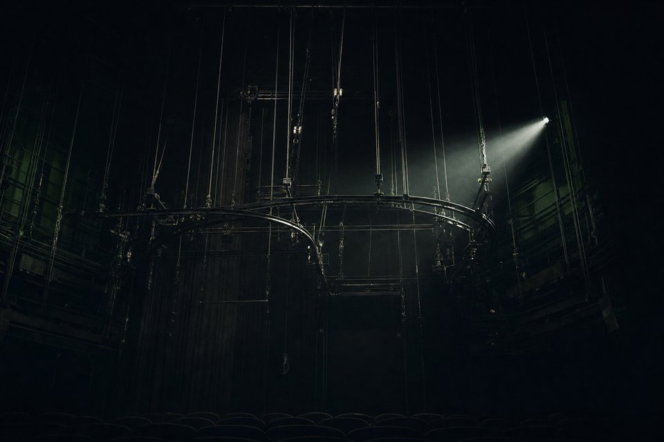 Aerial stage rigging and flying-harness track suspended above an empty Broadway theater, lit by a single spotlight